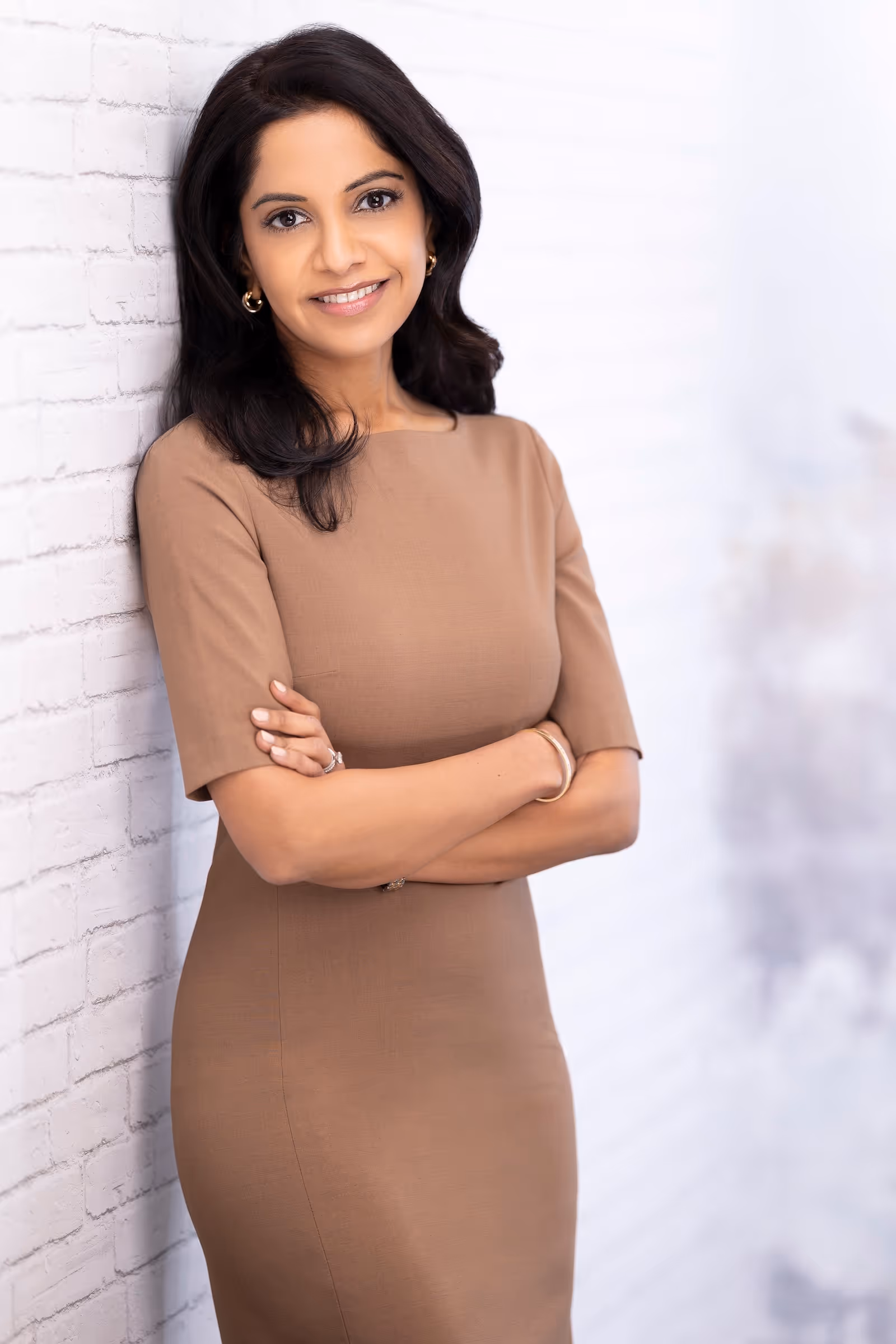 Dr. Shah smiling in a brown dress while leaning on a white wall