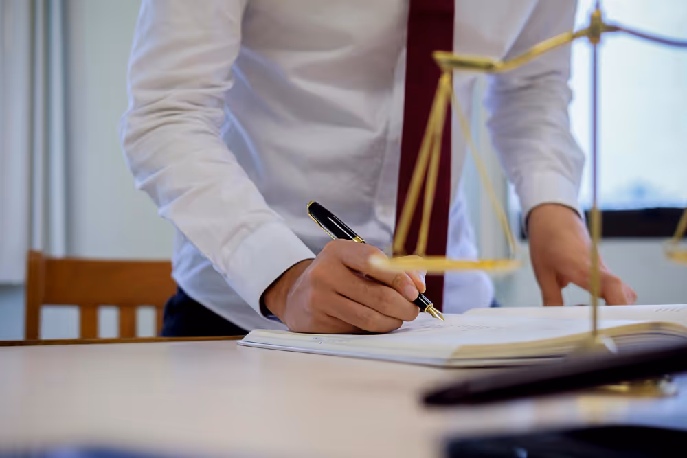 Person in weißem Hemd und roter Krawatte, der auf einem Schreibtisch ein Dokument mit einem Stift unterschreibt, daneben eine goldene Waage.