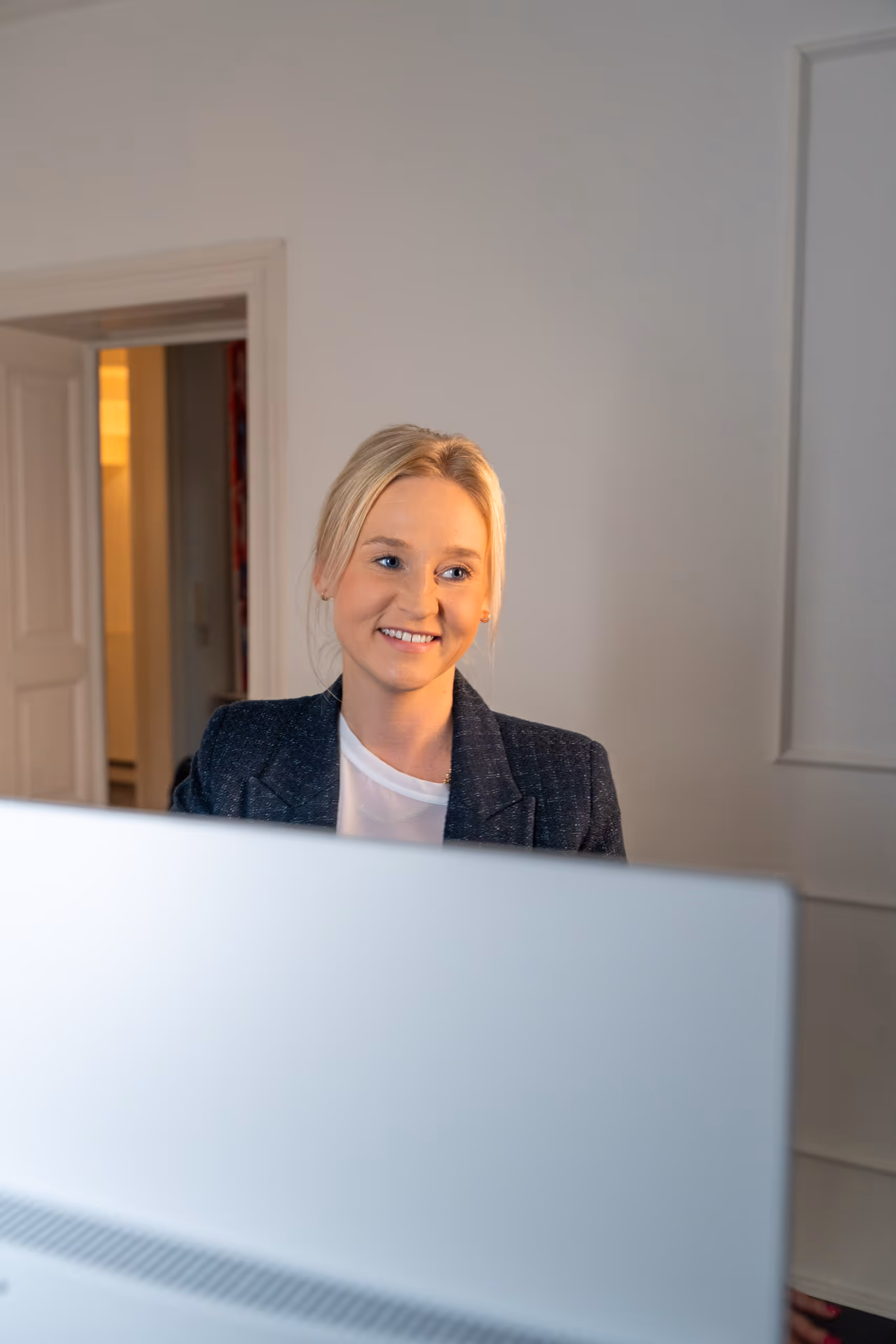 Lächelnde blonde Frau trägt Blazer und sitzt vor einem Computerbildschirm in einem Büro.