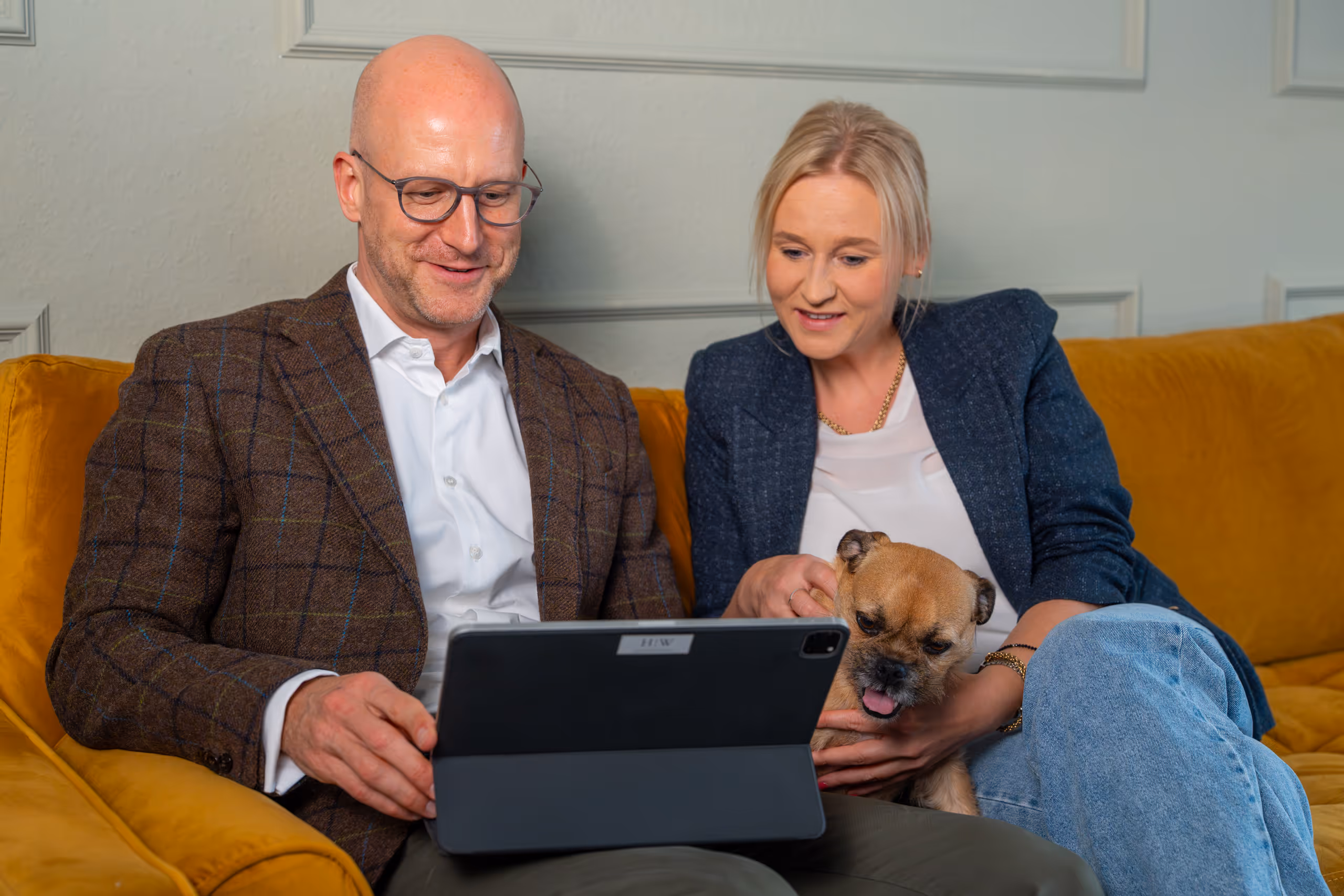 Ein Mann und eine Frau sitzen auf einem gelben Sofa, schauen gemeinsam auf ein Tablet, während die Frau einen kleinen Hund hält.