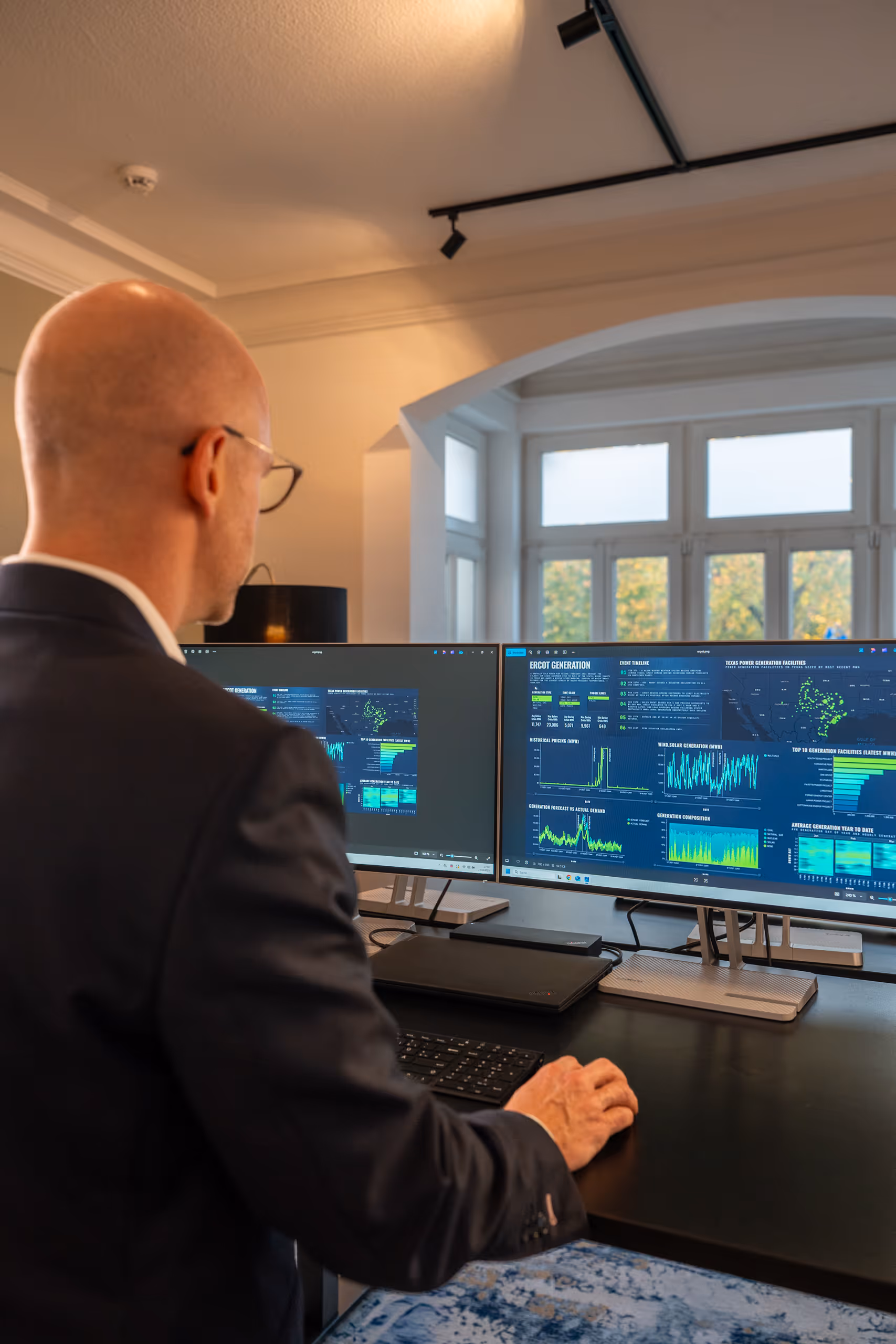 Mann im Anzug vor zwei Monitoren, die Daten und Diagramme anzeigen, in einem modernen Büro mit großen Fenstern.