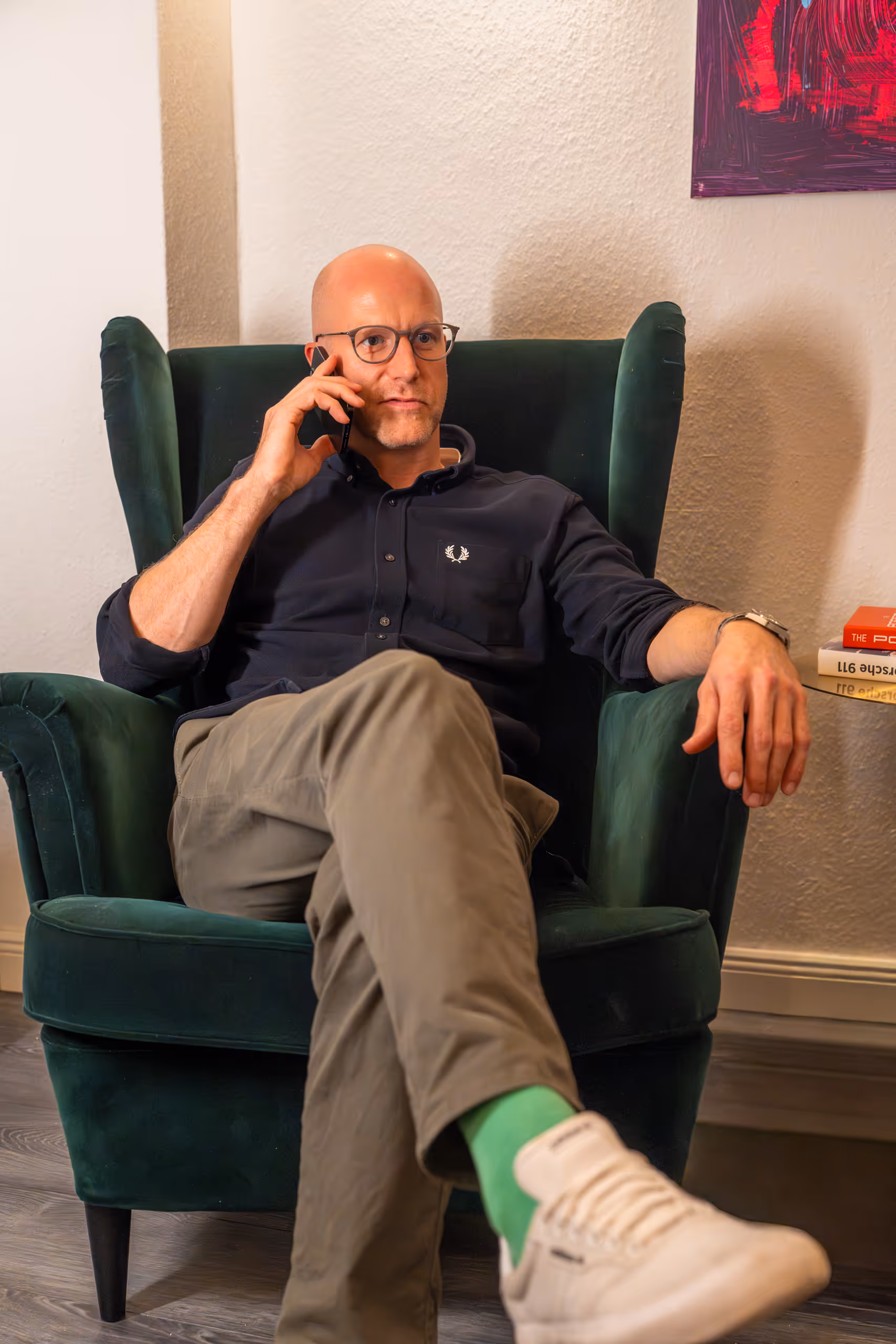 Ein Mann mit Brille sitzt in einem grünen Sessel, trägt khakifarbene Hose, grünen Socken und weiße Sneakers und spricht am Handy.