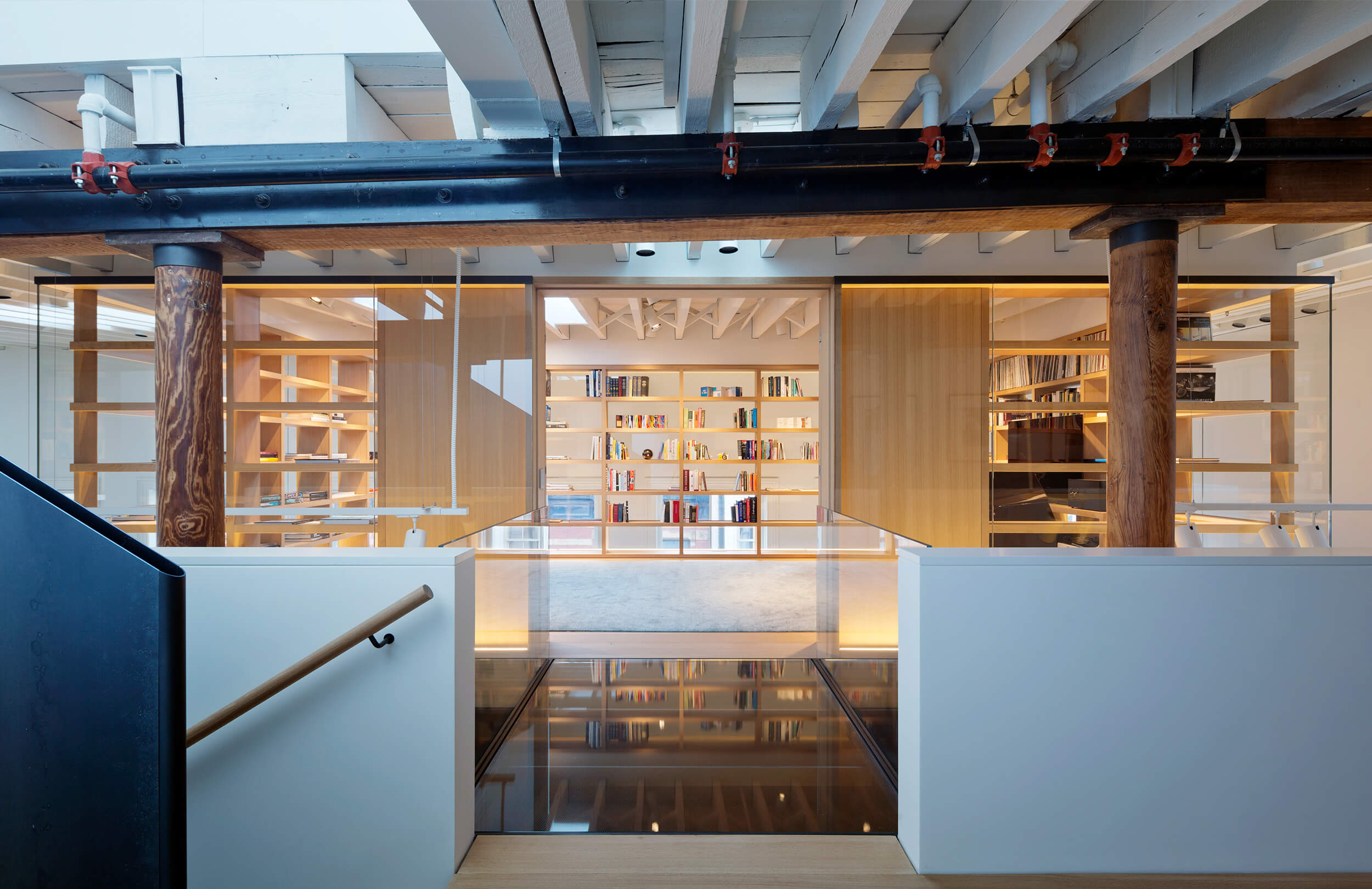 CNR Group NYC luxury home renovation: Modern loft interior with wooden shelves filled with books, exposed wooden beams, glass railing, and a descending staircase with a wooden handrail.
