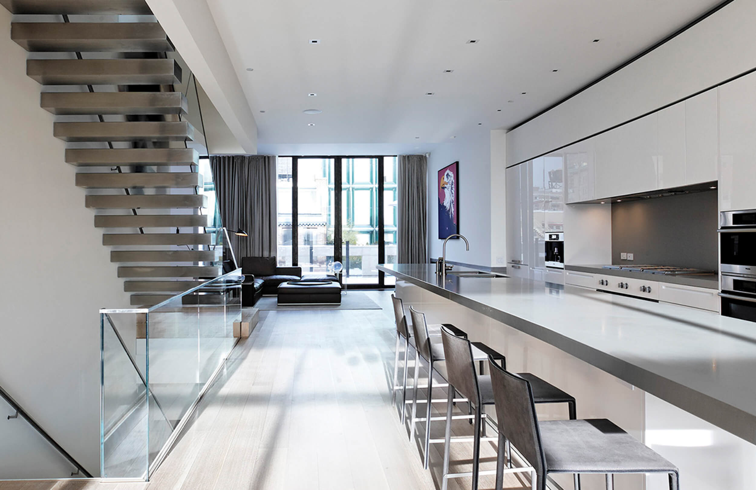 CNR Group NYC luxury home renovation: Modern kitchen with a long white island, four bar stools, stainless steel appliances, floating staircase, and large windows with curtains.