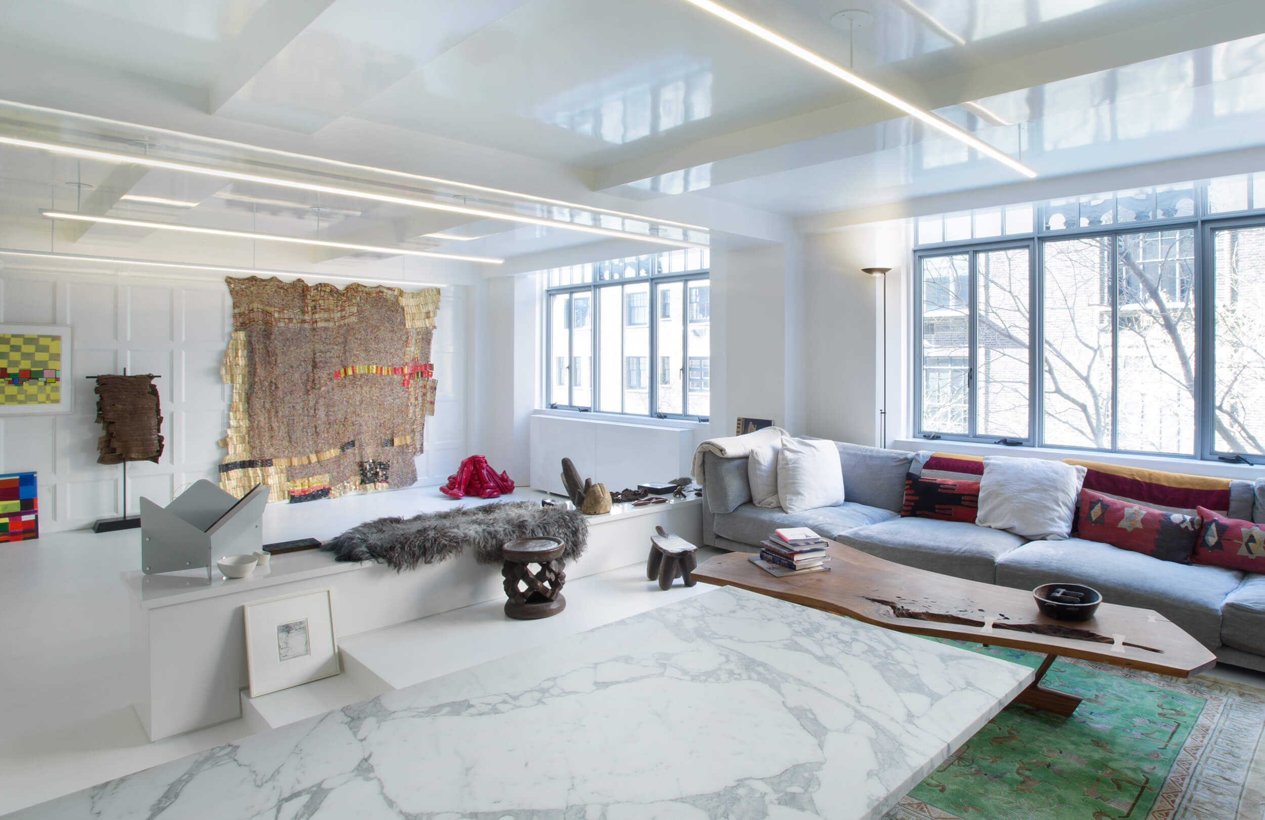 CNR Group NYC luxury home renovation: Bright modern living room with large windows, a gray sectional sofa with colorful pillows, wooden and marble coffee tables, and eclectic wall art.