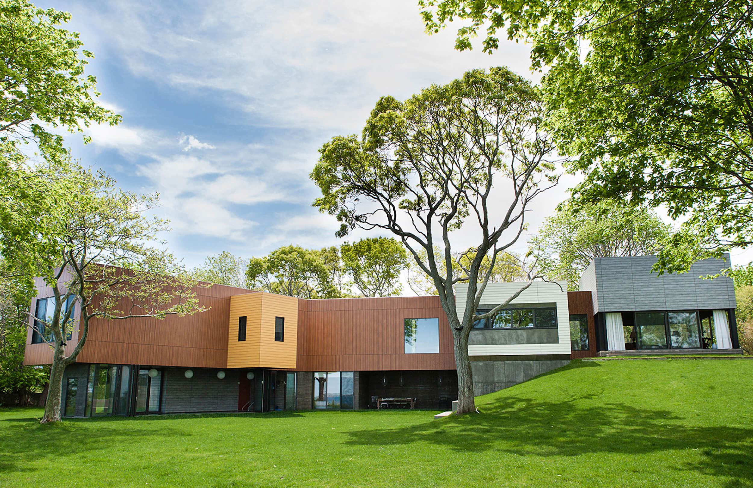 CNR Group luxury home construction: Modern multi-colored beach house with large windows surrounded by green lawn and trees under a partly cloudy sky.