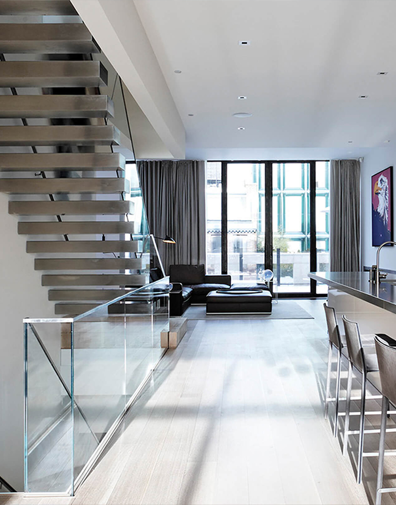 CNR Group NYC luxury home construction project: Modern open living space with floating staircase, glass railing, black sectional sofa, bar stools, and large floor-to-ceiling windows.