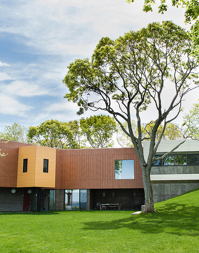 CNR Group luxury home construction project: Modern two-story house with wooden paneling and large windows, featuring a green lawn and a large tree in the foreground.