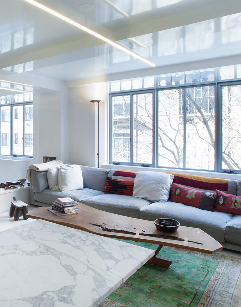 CNR Group NYC luxury home construction project: Modern living room with large gray sectional sofa, colorful pillows, wood coffee table, marble table, green patterned rug, and large windows.