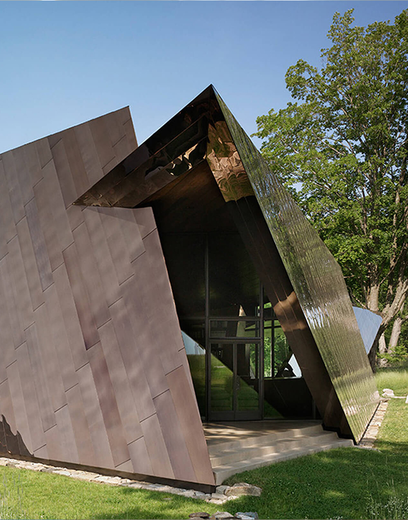 CNR Group luxury home construction project: Modern angular building with bronze and reflective metal panels, surrounded by grass and trees under a clear sky.