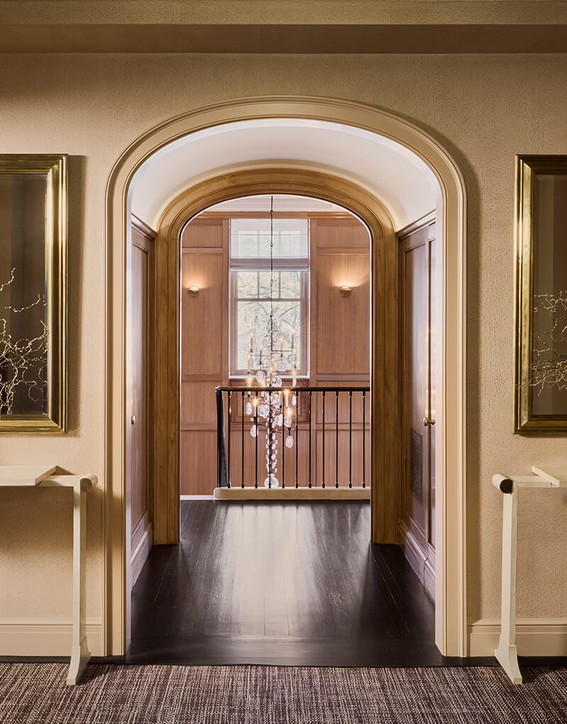CNR Group luxury home construction project: View through an arched doorway into a hallway with dark wood floors, wood-paneled walls, a window, and a hanging crystal chandelier.