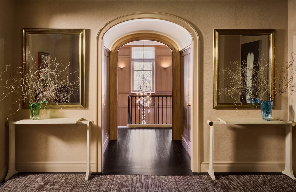 CNR Group luxury home builder project: Hallway with an arched doorway leading to a wooden paneled room and a crystal chandelier, flanked by two white console tables each holding a vase with decorative twigs beneath framed mirrors.