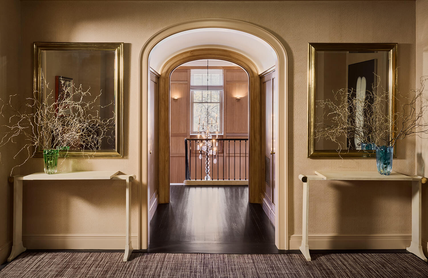 CNR Group luxury home builder project: Symmetrical hallway with arched doorway, two cream tables each holding a vase with dry branches, and large gold-framed mirrors above them.