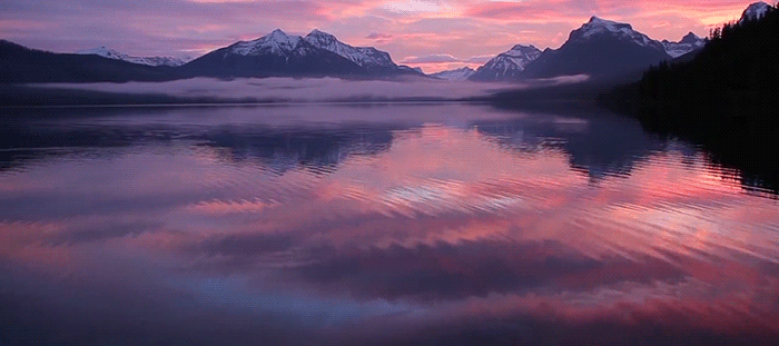 Calm lake at sunset reflecting snow-capped mountains and pink-purple clouds.
