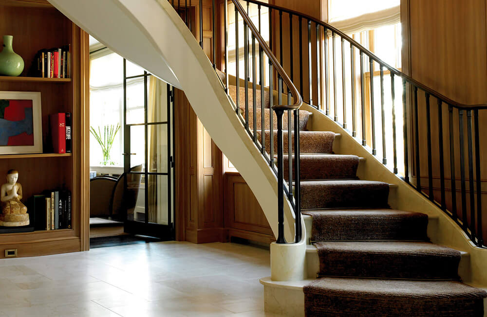 Curved staircase with carpet runner and black metalwork railing in a wood-paneled room with built-in bookshelf and glass door.