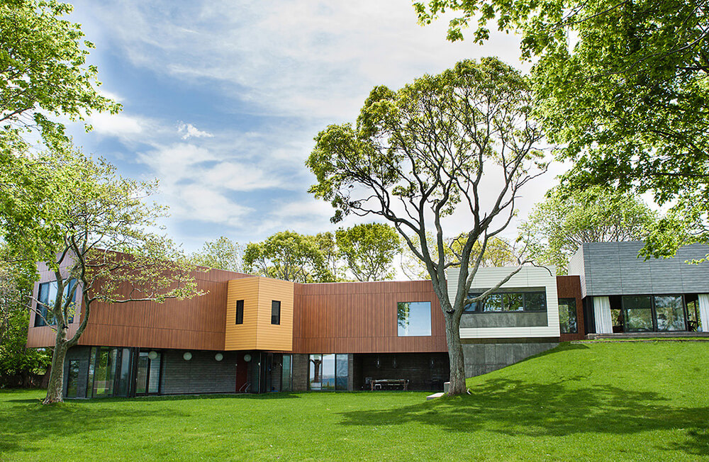 CNR Group luxury home builder project: Modern multi-section house with wood and concrete exterior surrounded by green grass and leafy trees under a partly cloudy sky.