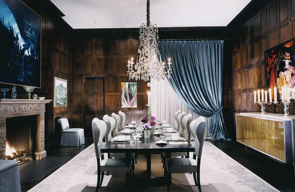 CNR Group NYC luxury home builder project interior: Elegant dining room with dark wood paneling, a long table set for twelve, crystal chandelier, fireplace, and blue drapes.