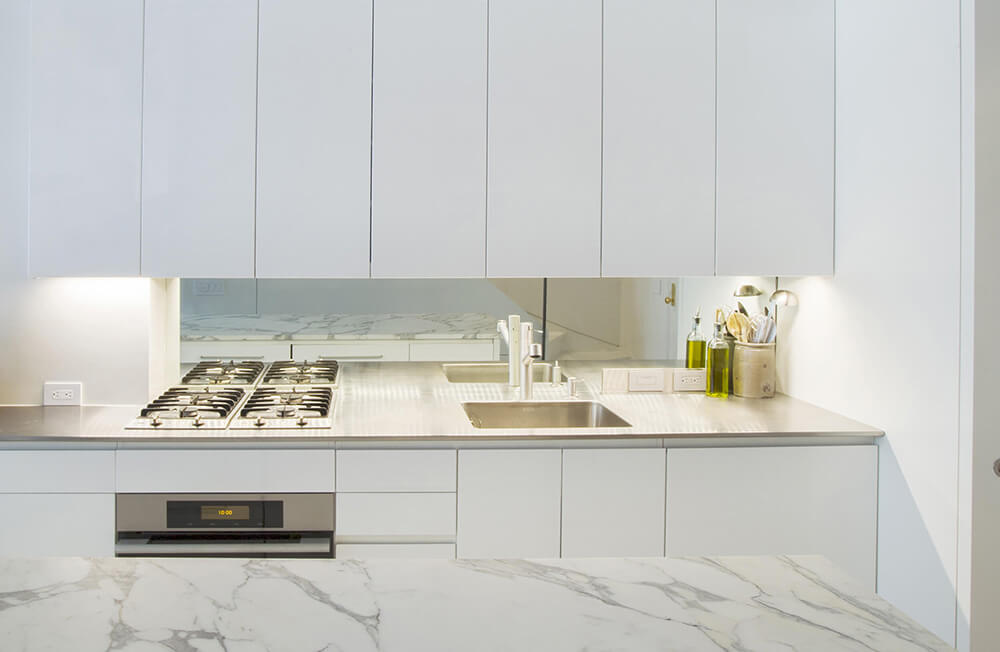 Minimalist white kitchen with gas stove, stainless steel sink, marble countertop island, and overhead custom millwork white cabinets.