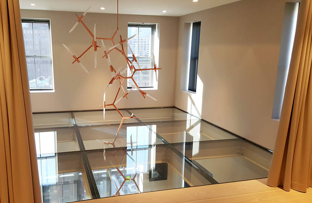 Modern room with large windows, beige curtains, and a unique hanging sculpture reflected on glass floor panels.