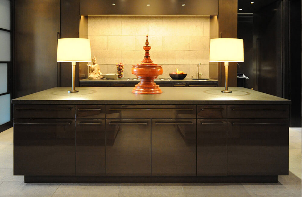 CNR Group NYC luxury home builder project interior: Modern kitchen island with two lit table lamps and a central decorative wooden urn, dark cabinetry, and a beige tiled backsplash.