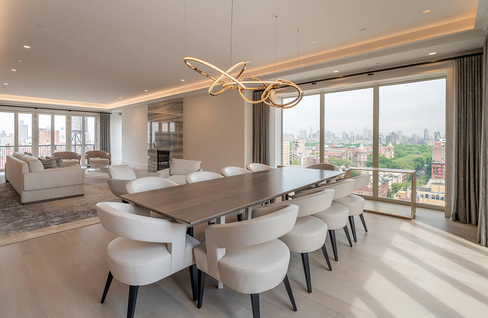 CNR Group NYC luxury home builder project interior: Modern dining room with a long wooden table surrounded by white upholstered chairs and large windows overlooking a cityscape with greenery.