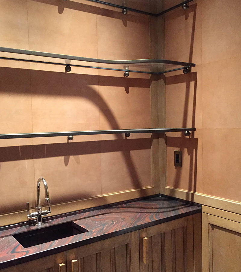 Corner kitchen countertop with a mounted silver faucet, two glass shelves, and custom millwork wood paneled cabinetry below.