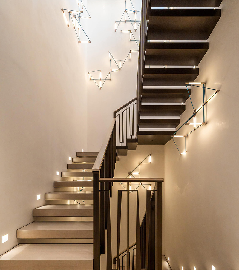 Modern staircase with floating wooden steps, black metalwork railing, and geometric wall-mounted light fixtures.