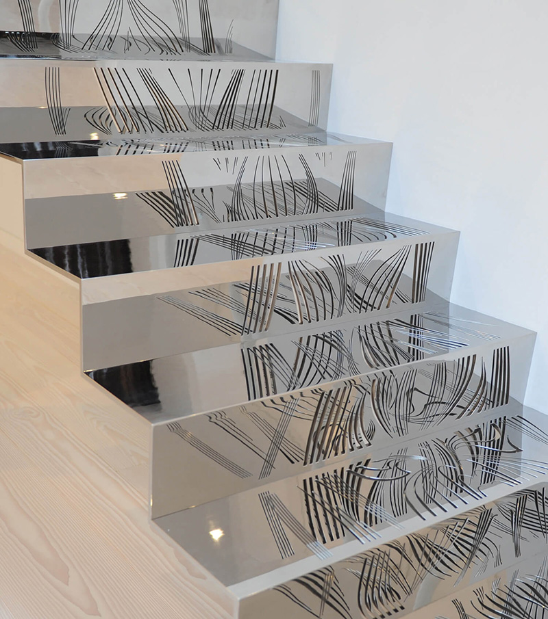 Shiny metalwork staircase with black abstract line patterns reflected on each step, next to a light wood floor and white wall.