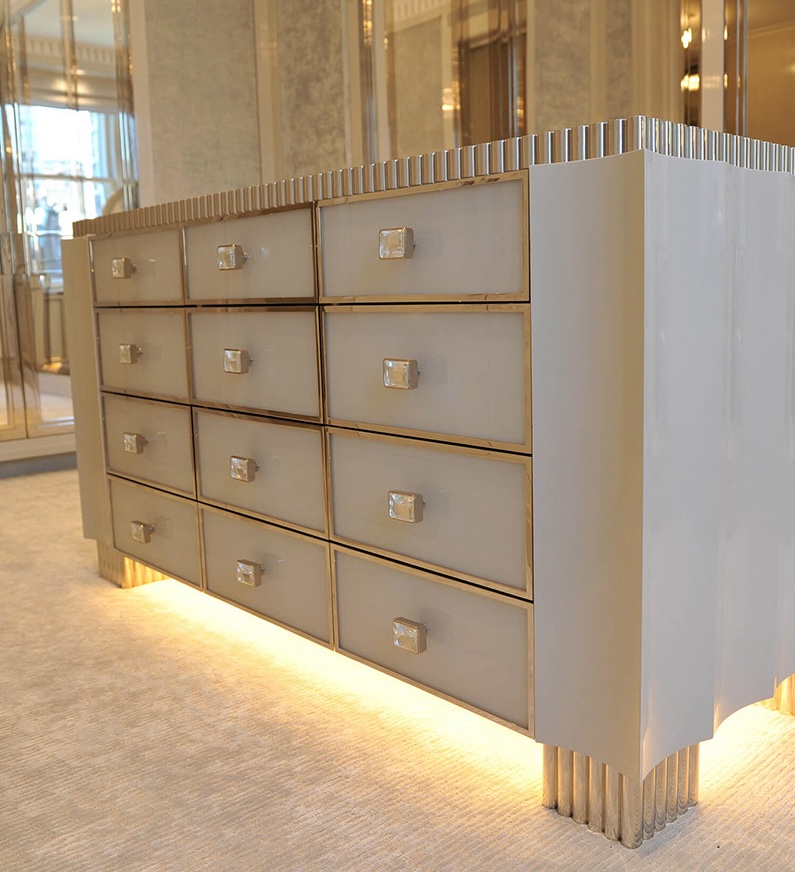 Modern white and gold custom millwork dresser with twelve drawers and illuminated base standing on a beige carpet.