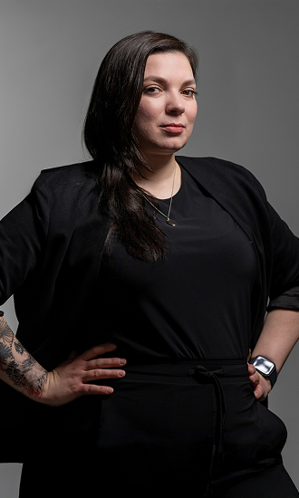CNR Group luxury builders team member: Patrycja. Confident woman with long dark hair wearing black clothing, a necklace, and a smartwatch, standing with hands on hips against a gray background.