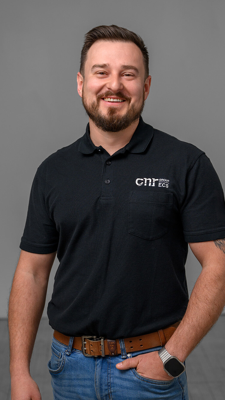 CNR Group luxury builders team member: Dawid. Smiling man with short brown hair and beard wearing a black polo shirt with logo, blue jeans, brown belt, and smartwatch with one hand in pocket.