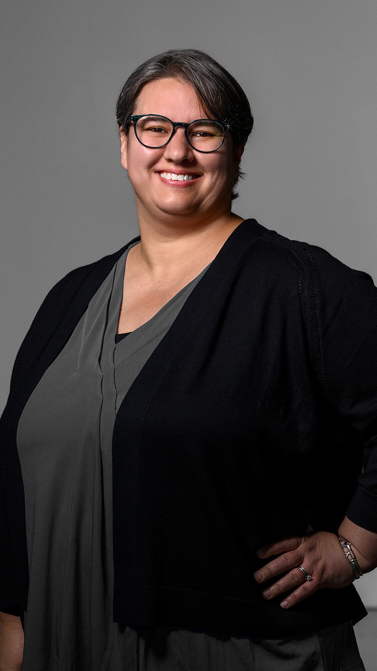 CNR Group luxury builders team member: Ashley. Smiling person with short dark hair and glasses wearing a dark cardigan over a grey top, standing against a plain grey background.