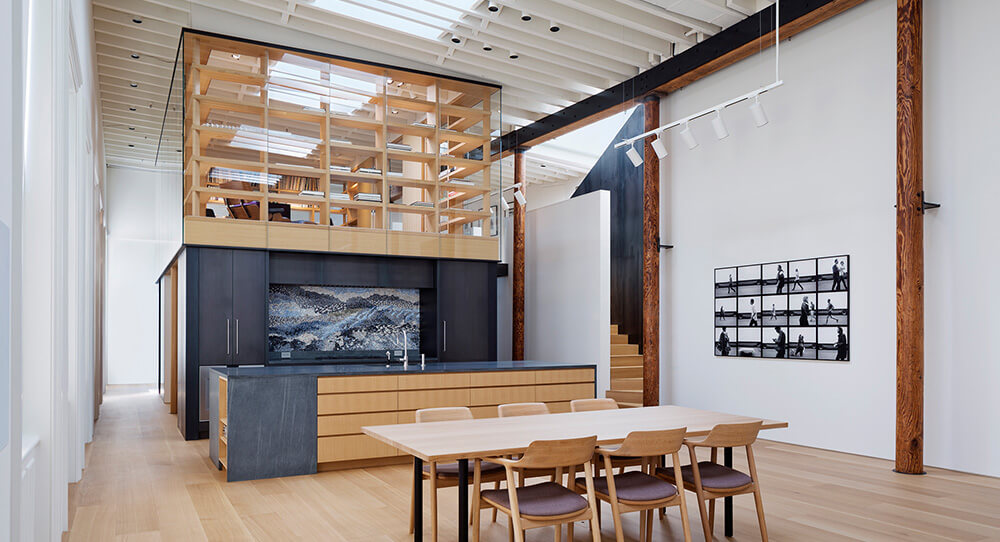 CNR Group NYC luxury builders: Modern open-concept kitchen and dining area with light wood floor, a central island with black countertop, and a glass-enclosed upper shelving unit with custom millwork wooden racks.