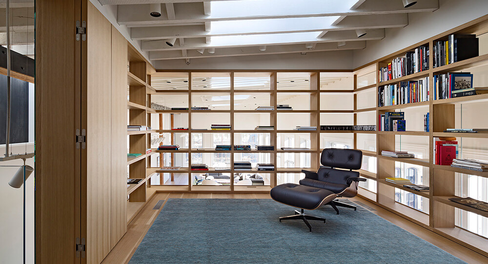 CNR Group NYC luxury builders: Modern reading nook with glass walls, wooden shelves filled with books, a blue rug, and a black leather lounge chair and ottoman.