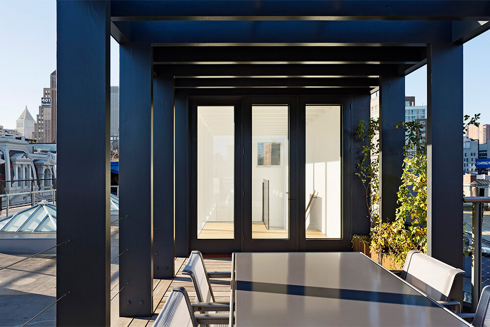 CNR Group NYC luxury builders: Modern rooftop patio with black metal pergola, glass table, metal chairs, and potted plants, overlooking city buildings.