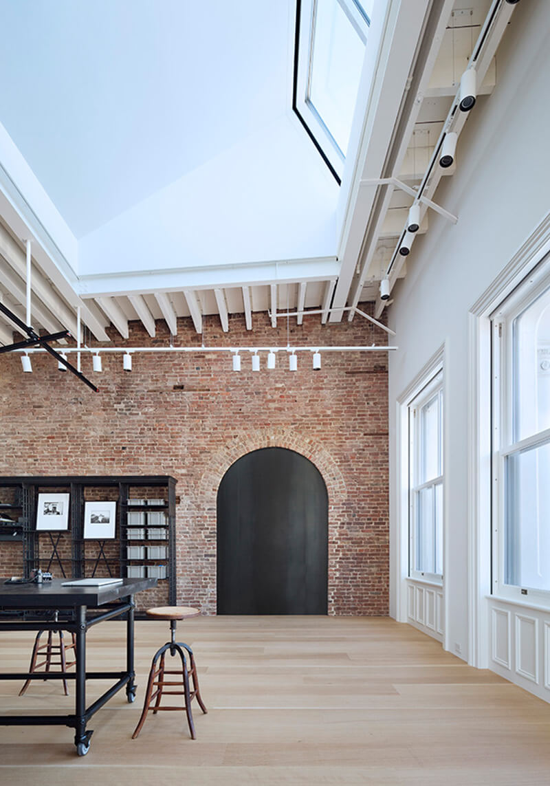CNR Group NYC luxury builders: Spacious modern room with a high ceiling skylight, exposed brick wall, arched black door, wooden floor, and industrial-style black furniture and stools.