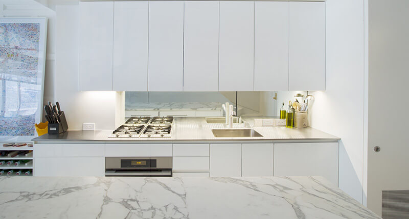 CNR Group luxury home project management: Minimalist white kitchen with marble countertop, stainless steel sink, gas stove, and white custom millwork cabinets with a mirrored backsplash.