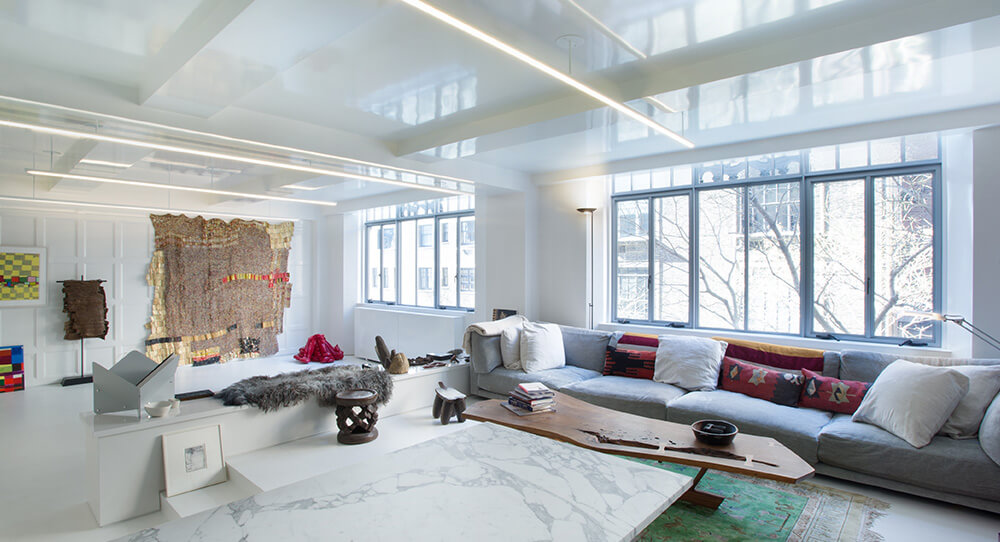 CNR Group NYC luxury home builders: Bright, modern living room with large windows, a long gray sectional sofa with colorful pillows, a low wooden coffee table, and decorative art pieces on white walls.