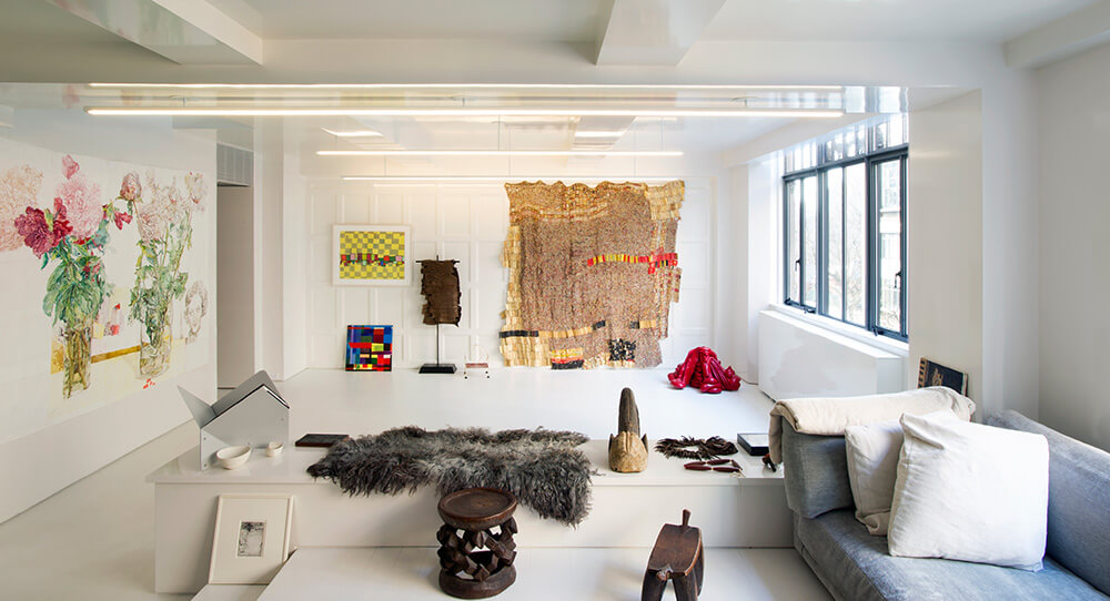 CNR Group NYC luxury home project management: Bright modern living space with white walls, large windows, contemporary art on walls, grey sofa with white pillows, and decorative objects on a white platform.