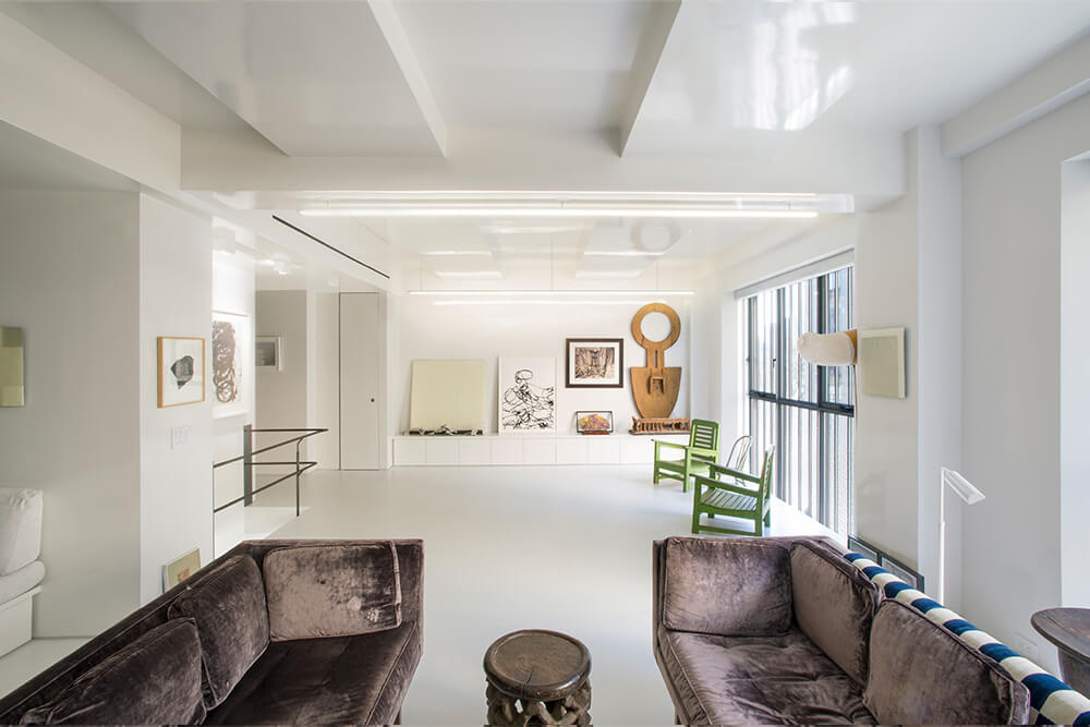 CNR Group NYC luxury home project management: Bright modern living room with white walls, two dark velvet sofas, a small round wooden table, green wooden chairs, and artwork displayed on the far wall.