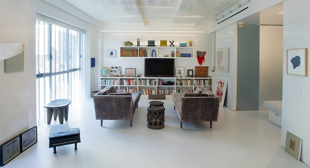 CNR Group NYC luxury home project management: Modern living room with two gray velvet sofas facing a wall-mounted TV above bookshelves, featuring decorative items and large windows on the left.
