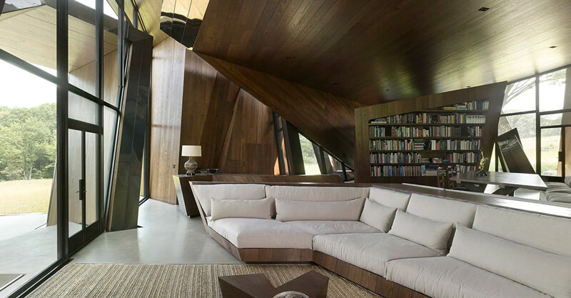 CNR Group luxury home construction: Modern living room with large gray sectional sofa, wooden bookshelf, slanted wooden ceiling, and floor-to-ceiling glass doors opening to an outdoor area.