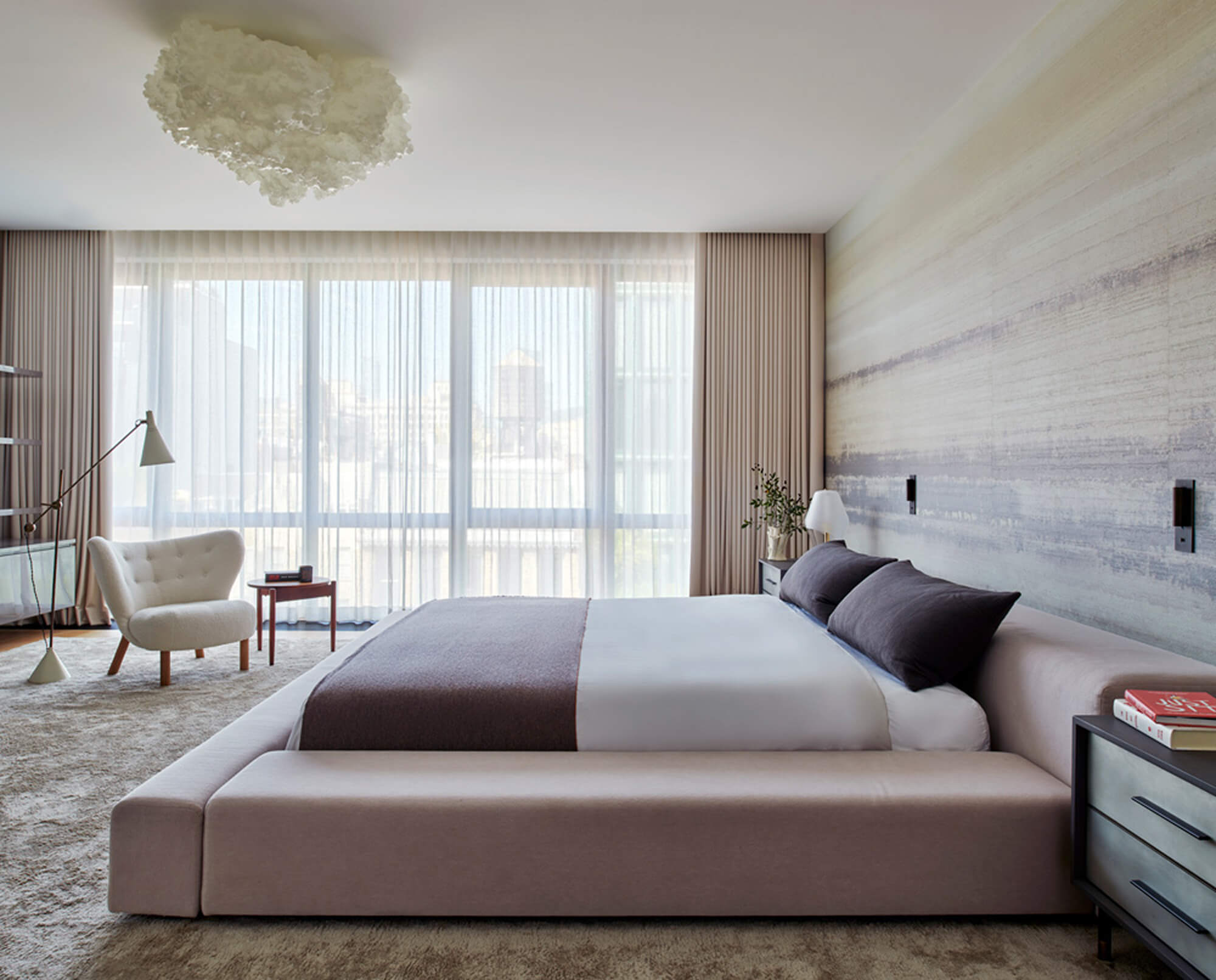 CNR Group luxury home project management: Modern bedroom with a low-profile upholstered bed, dark pillows, beige throw, large window with sheer curtains, and a white armchair with floor lamp.