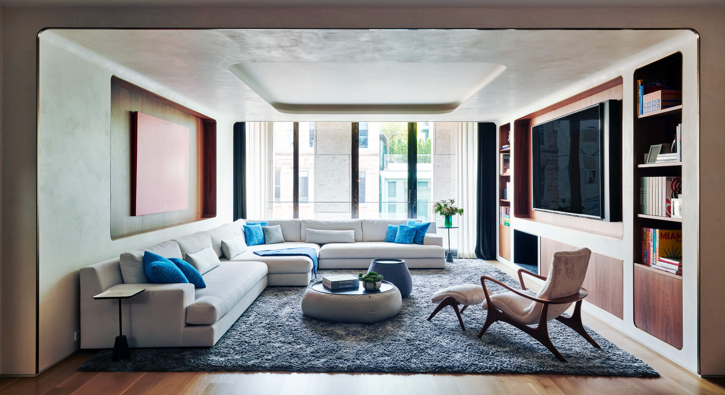 CNR Group NYC luxury home project management: Modern living room with large white sectional sofa, blue pillows, shaggy gray rug, wooden lounge chair with ottoman, built-in bookshelves, and large windows.