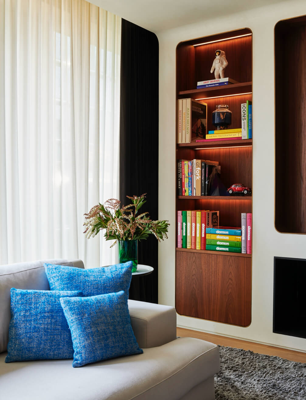 CNR Group NYC luxury home project management: Modern living room corner with a light gray sofa adorned with blue cushions, a side table with a green vase of flowers, sheer white and black curtains, and a built-in wooden bookshelf with books, a small astronaut figurine, and a miniature red car.