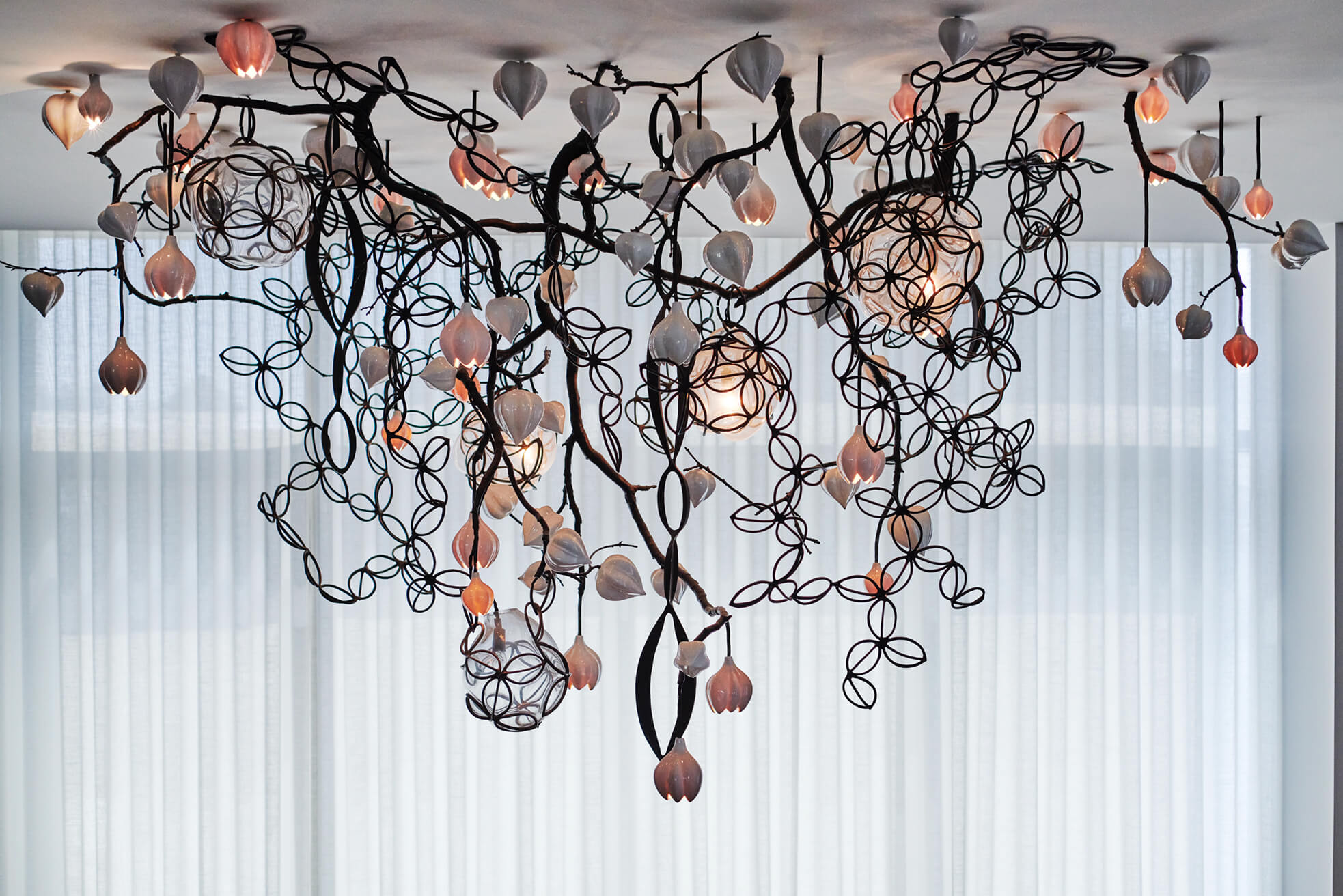 Intricate custom metalworks black metal chandelier with floral patterns and soft pink glass bulbs hanging from the ceiling in front of sheer white curtains.