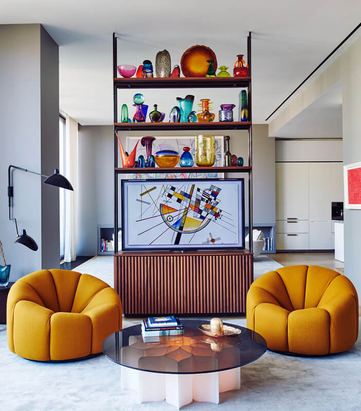 CNR Group NYC luxury home project management: Modern living room with two mustard yellow rounded armchairs, a round glass coffee table, and a wooden shelving unit displaying colorful glass vases and abstract artwork.
