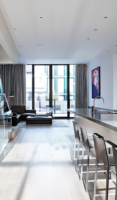 CNR Group luxury home construction interior: Modern living area with black leather sofa, large glass doors with grey curtains, long kitchen counter with tall chairs, and a colorful painting on the wall.