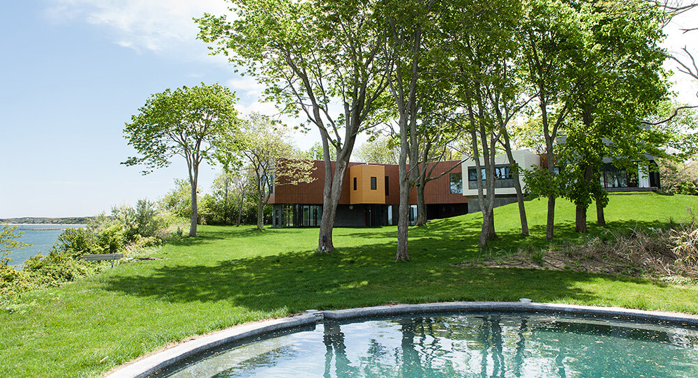 CNR Group luxury home construction: Modern house with large windows behind trees on a grassy hill near a water body and a circular pool in the foreground.