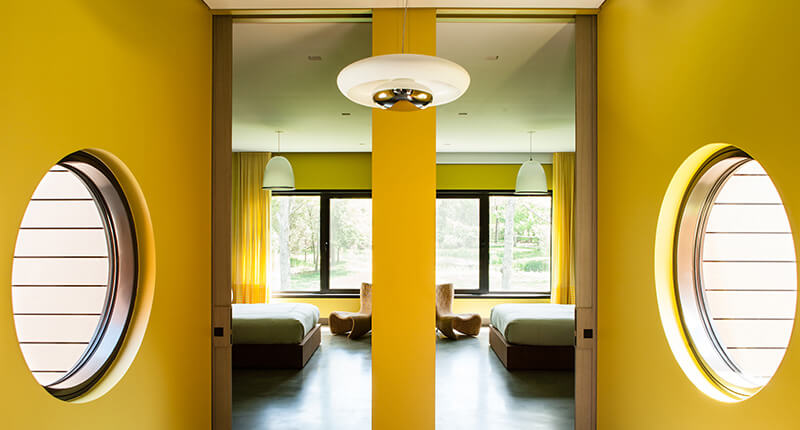 CNR Group luxury home construction interior: Modern bedroom with yellow walls, round windows, two beds, and a central mirrored column reflecting the room.